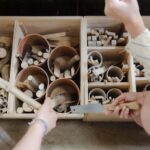 Ein Bild von zwei Kisten mit Unterteilungen und vielen unterschiedlichen Holzstücken darin. Im Vordergrund sieht man Hände die Holz und eine Säge halte.