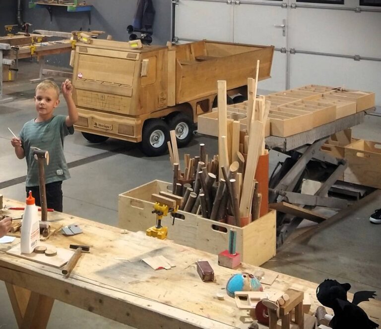 Kind steht vor Konrad dem Holz LKW, hinter einer Werkbank in einer Halle und winkt
